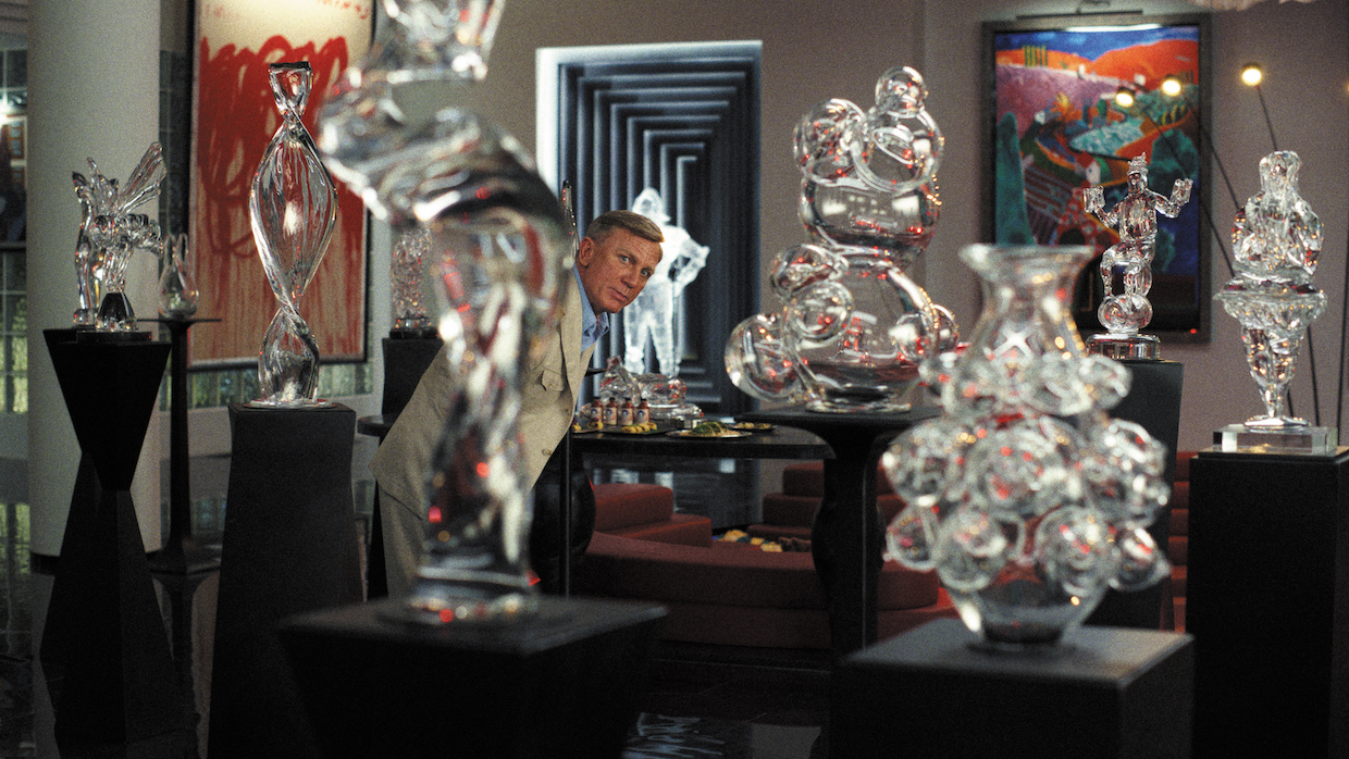 Detective Benoit Blanc (Daniel Craig) stands in a room surrounded by glass figurines on black pedestals.