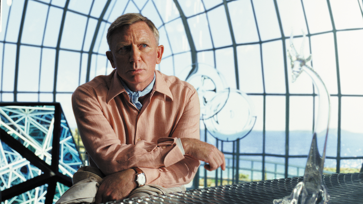 Detective Benoit Blanc, played by Daniel Craig, sits in a glass atrium.