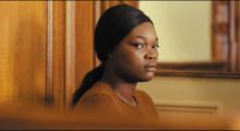A Black woman sits in a courtroom, wearing a brown sweater and straight black hair pulled back in a low ponytail.
