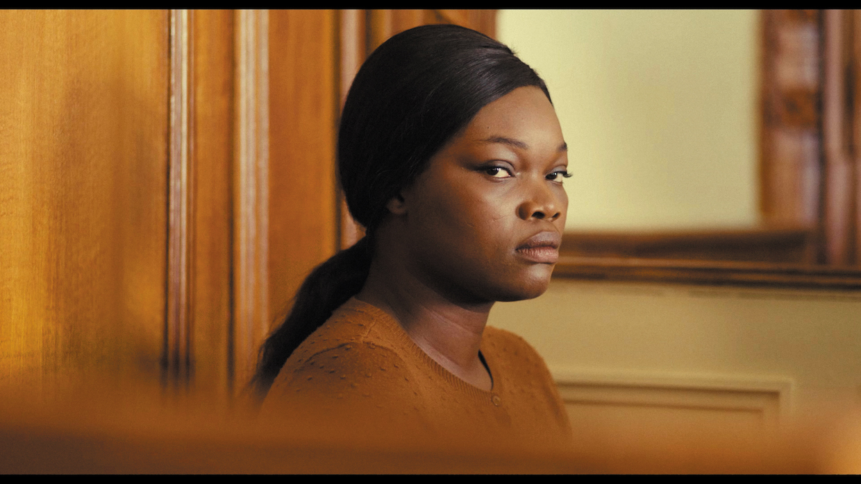 A Black woman sits in a courtroom, wearing a brown sweater and straight black hair pulled back in a low ponytail.