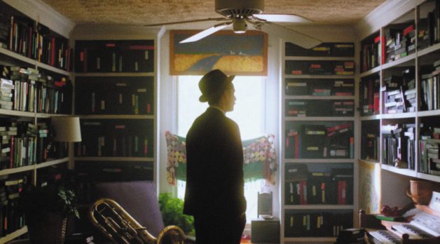A man wearing a suit and top hat stands in front of walls lined with built-in bookcases, each shelf filled with VHS tapes.