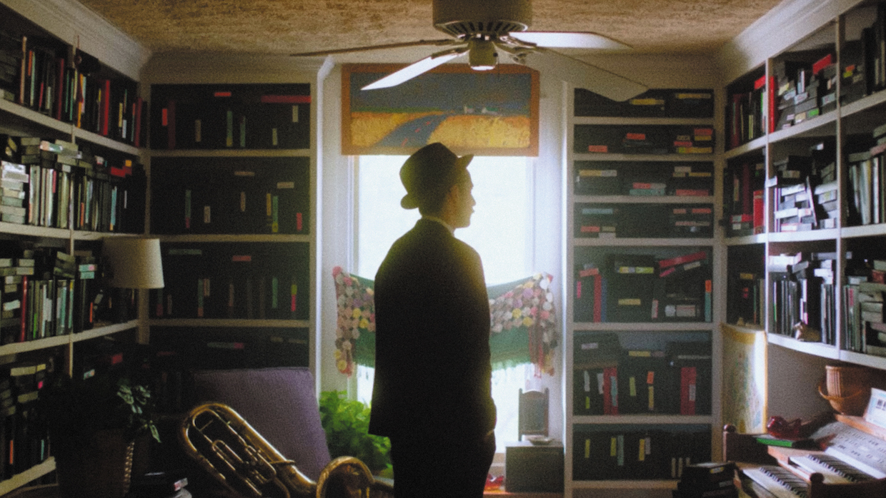 A man wearing a suit and top hat stands in front of walls lined with built-in bookcases, each shelf filled with VHS tapes.