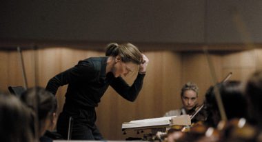 A blond woman conducts a string orchestra, wearing a black long-sleeve button up and slacks. She poses dramatically with her arm bent at the elbow and her fist pumped toward the air.