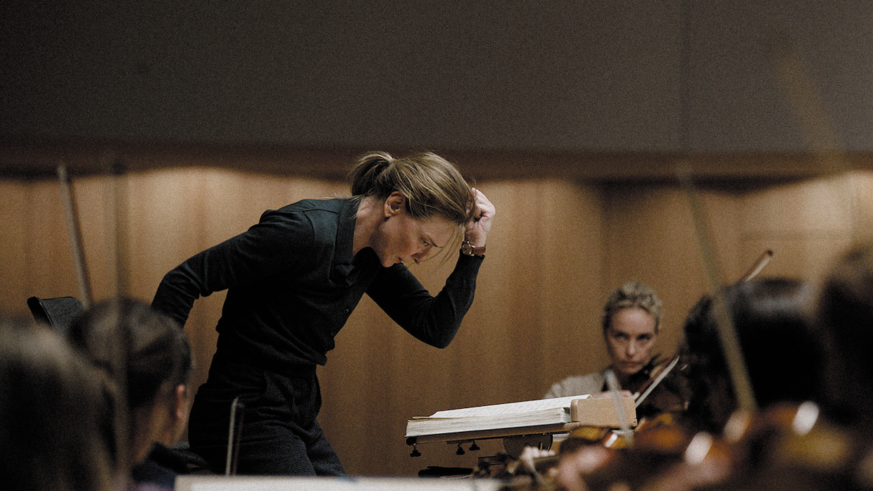 A blond woman conducts a string orchestra, wearing a black long-sleeve button up and slacks. She poses dramatically with her arm bent at the elbow and her fist pumped toward the air.