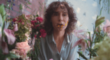 A woman with curly brown hair and a blue button-up blouse sits among a flower garden, orange carnation petals coming out of her mouth and sticking to her face.