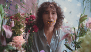 A woman with curly brown hair and a blue button-up blouse sits among a flower garden, orange carnation petals coming out of her mouth and sticking to her face.