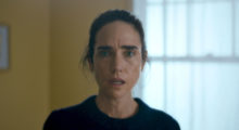 Actress Jennifer Connelly stands in the center of the frame, her hair tied back, wearing a black sweater. She is in a yellow-walled room with a window behind her. It is a dull environment, she looks perplexed.