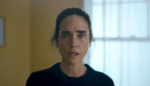 Actress Jennifer Connelly stands in the center of the frame, her hair tied back, wearing a black sweater. She is in a yellow-walled room with a window behind her. It is a dull environment, she looks perplexed.