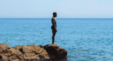 A Black woman wearing a black and blue one piece bathing suit stands on the edge of a cliff overlooking the deep blue sea.