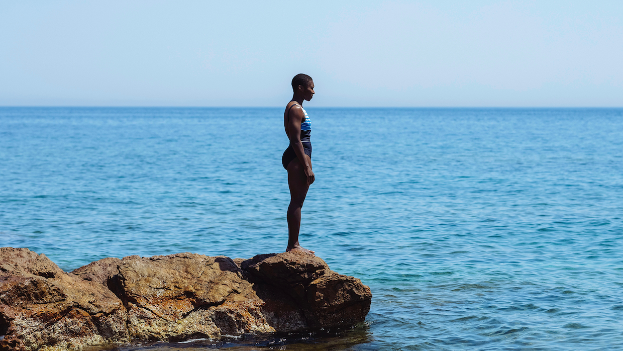 A Black woman wearing a black and blue one piece bathing suit stands on the edge of a cliff overlooking the deep blue sea.