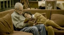 An elderly man with white hair, glasses, jeans and a gray sweater holds a book open and reads to his wife, with brown curly hair, a camel-colored cardigan and matching slacks. She leans on his shoulder as they sit on the couch.