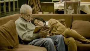 An elderly man with white hair, glasses, jeans and a gray sweater holds a book open and reads to his wife, with brown curly hair, a camel-colored cardigan and matching slacks. She leans on his shoulder as they sit on the couch.