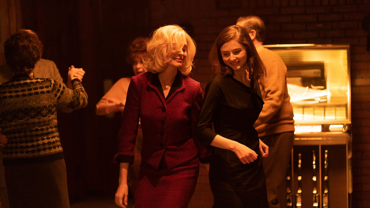 Two women, one blonde and one brunette, wearing 1960s-era outfits dance in front of a jukebox.