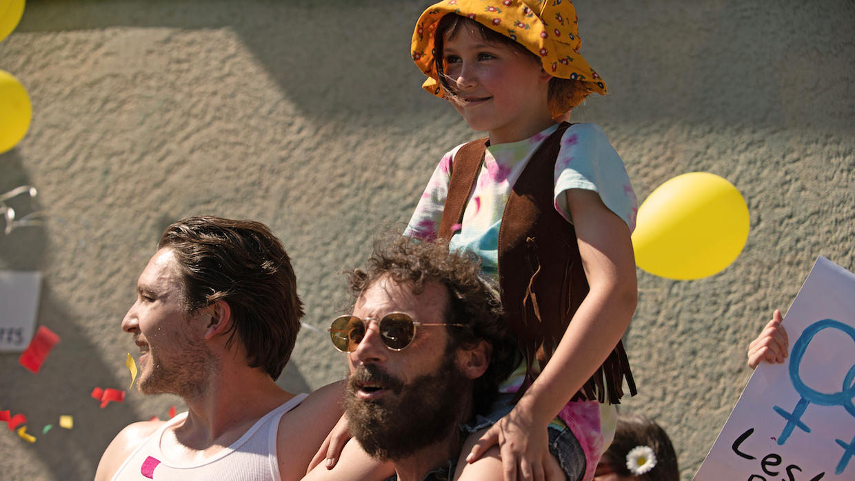 A young girl wearing a brown vest and bucket hat sits on her bearded father's shoulders as a parade passes them by.