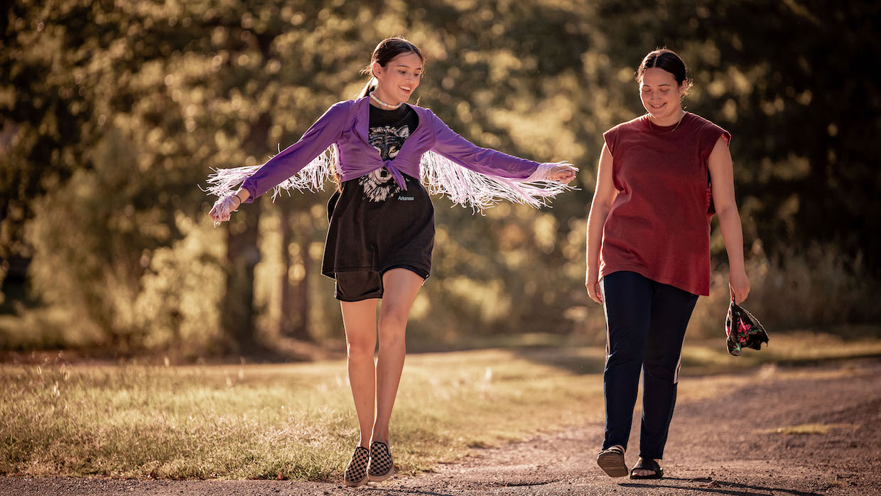 Two young women walk along a dirt path in a park. One wears a purple fringe jacket, a band t-shirt and black bike shorts with checkered vans. The other wears a sleeveless red t-shirt, black jeans and sandals.