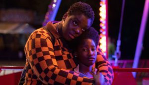 A Black mother and daughter sit in front of a carnival ride with purple light illuminating their faces.