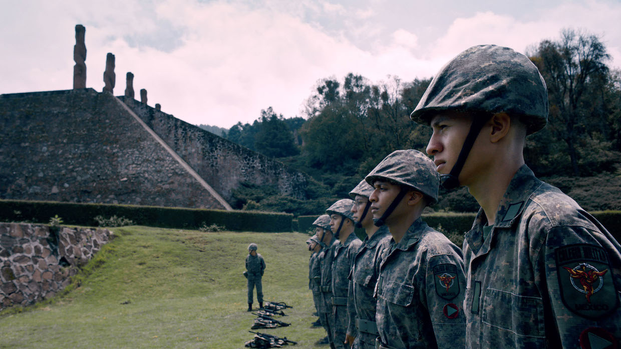 A line of Mexican soldiers in camouflage uniform stand in a neat line with an Aztec pyramid in the background.