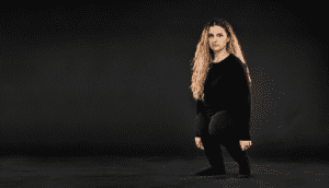 A woman with long, curly blond hair wears all black and stands amid a dark gray background. She has a rare condition where her legs are disproportionally short compared to her body.