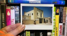 A polaroid photo of a stone building is held by a hand in front of various VHS tapes on a shelf, including David Cronenberg's Videodrome.