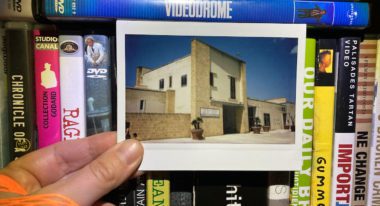 A polaroid photo of a stone building is held by a hand in front of various VHS tapes on a shelf, including David Cronenberg's Videodrome.