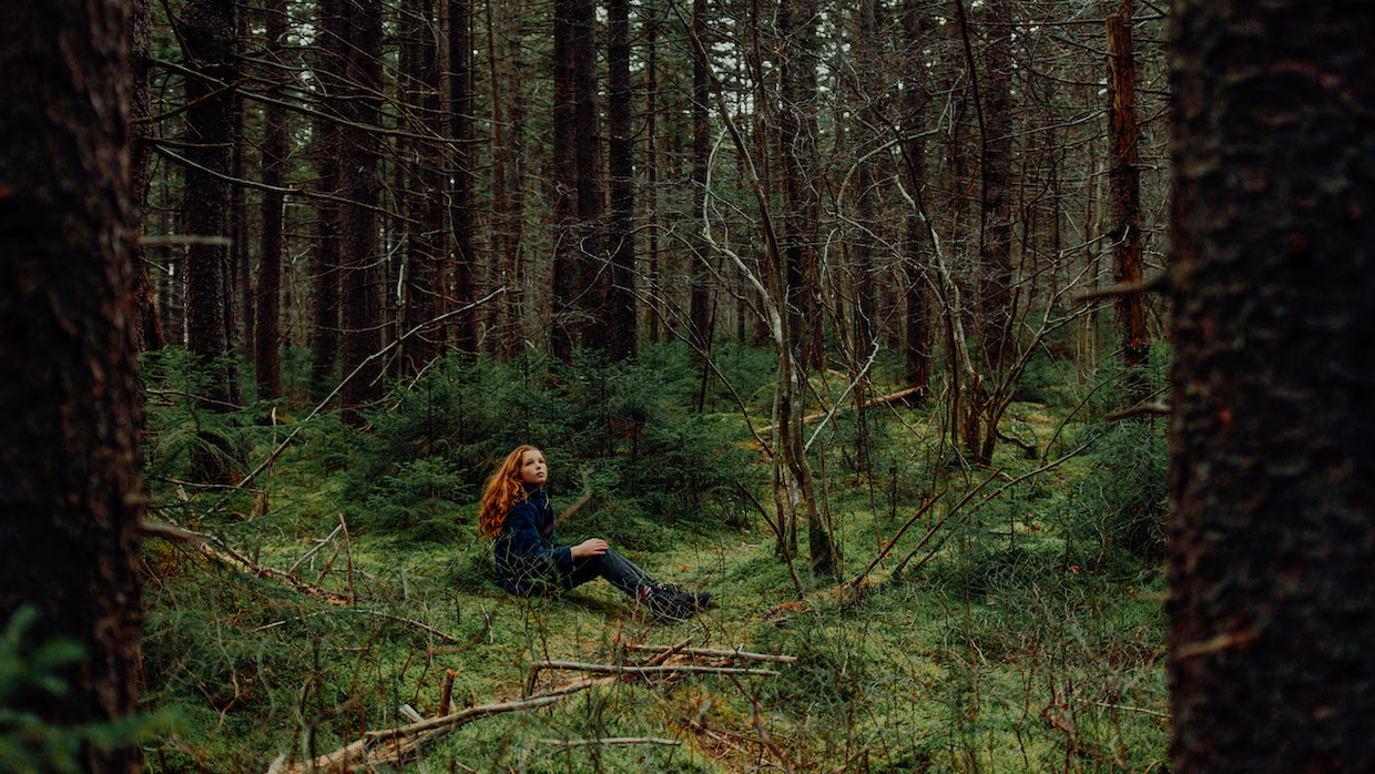 A woman with long, wavy red hair sits in a forrest surrounded by tall trees and lush grass.