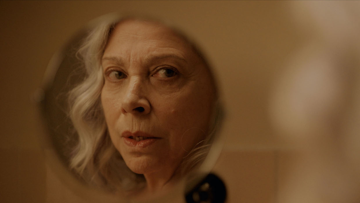 An older woman with gray hair looks at her reflection in a compact mirror.