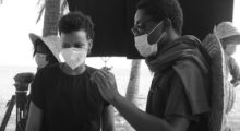 Two people wearing masks observe their surroundings on a film set, with other masked people roaming in the background. The image in in black and white.