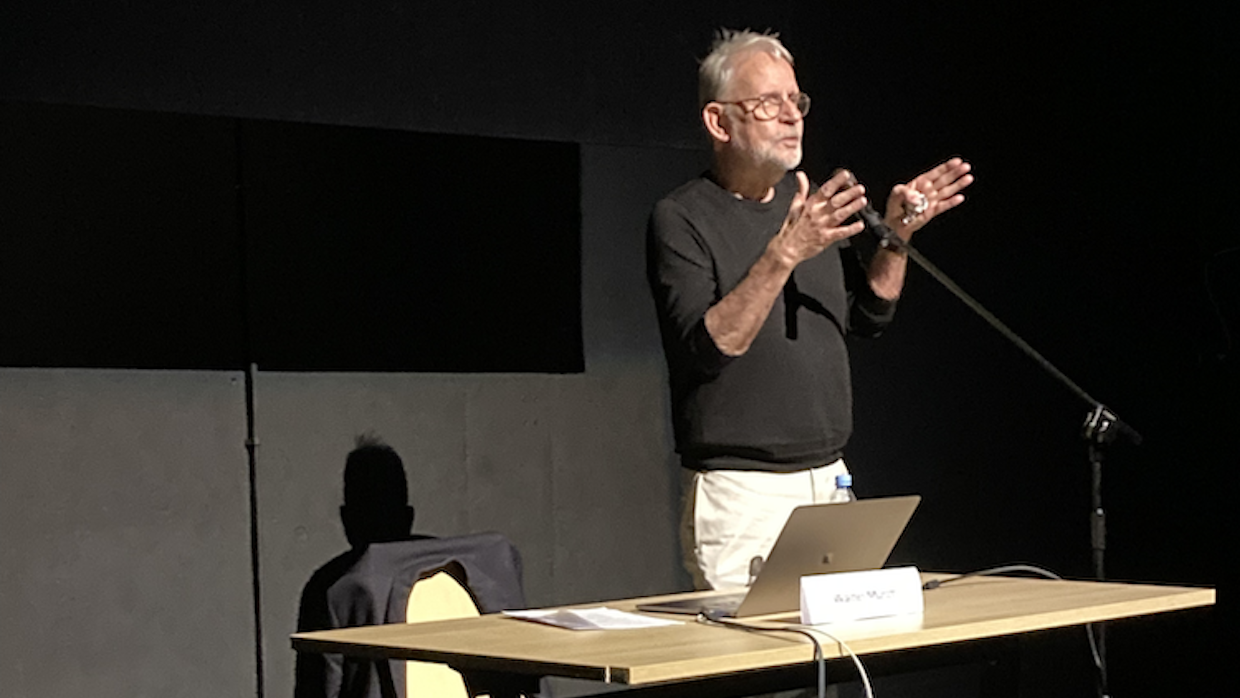 A man with white hair and a trimmed beard stands in front of a desk giving a lecture.