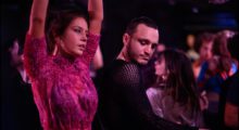 A man and a woman dance in a crowded club. She wears a fuchsia blouse and he ears a black long-sleeved mesh top.