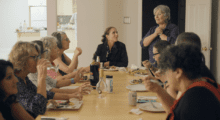 A group of women sit around a long dining table, eating food and drinking wine while having conversation.