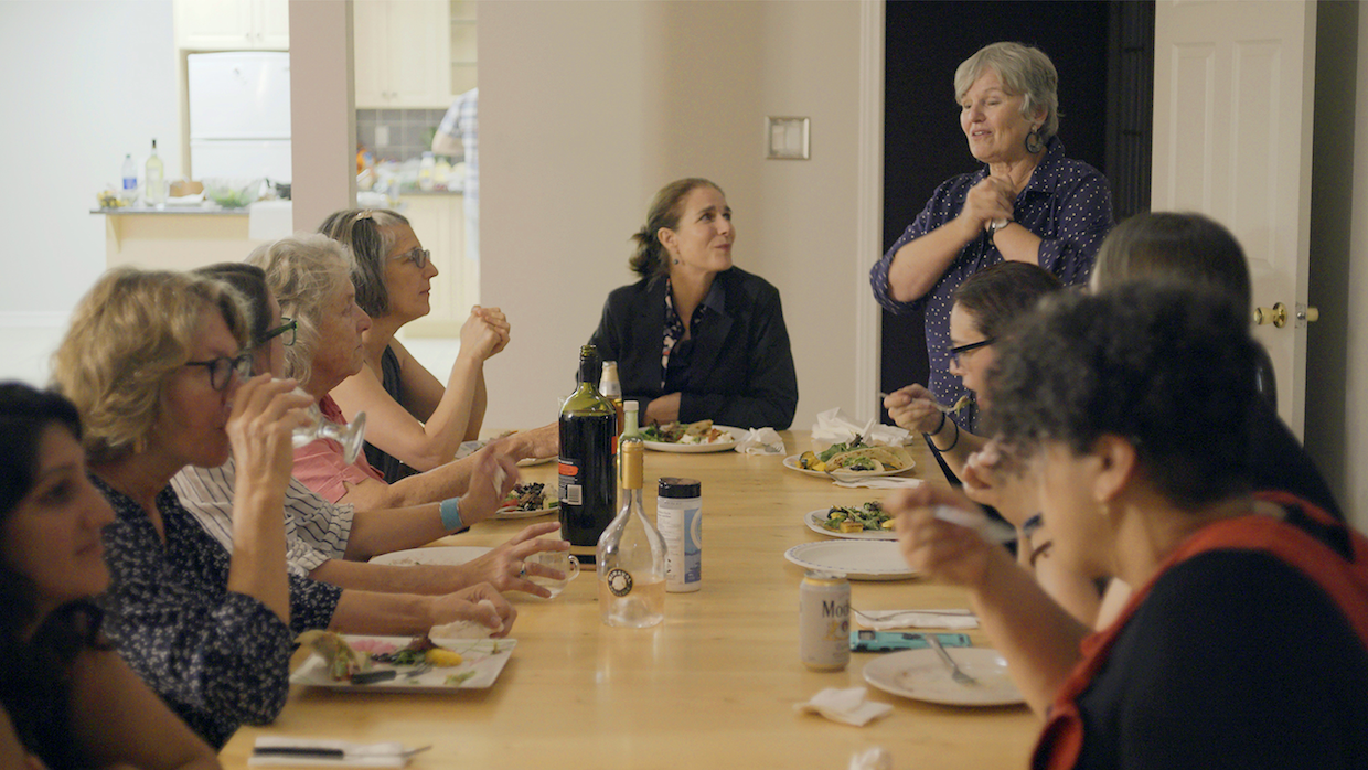 A group of women sit around a long dining table, eating food and drinking wine while having conversation.