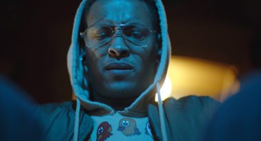 A young man wears a grey hoodie with the hood over his head and an unbottoned blue jacket. He also dons clear glasses frames. A blue light washes over him as he gazes down at something with his arms raised.