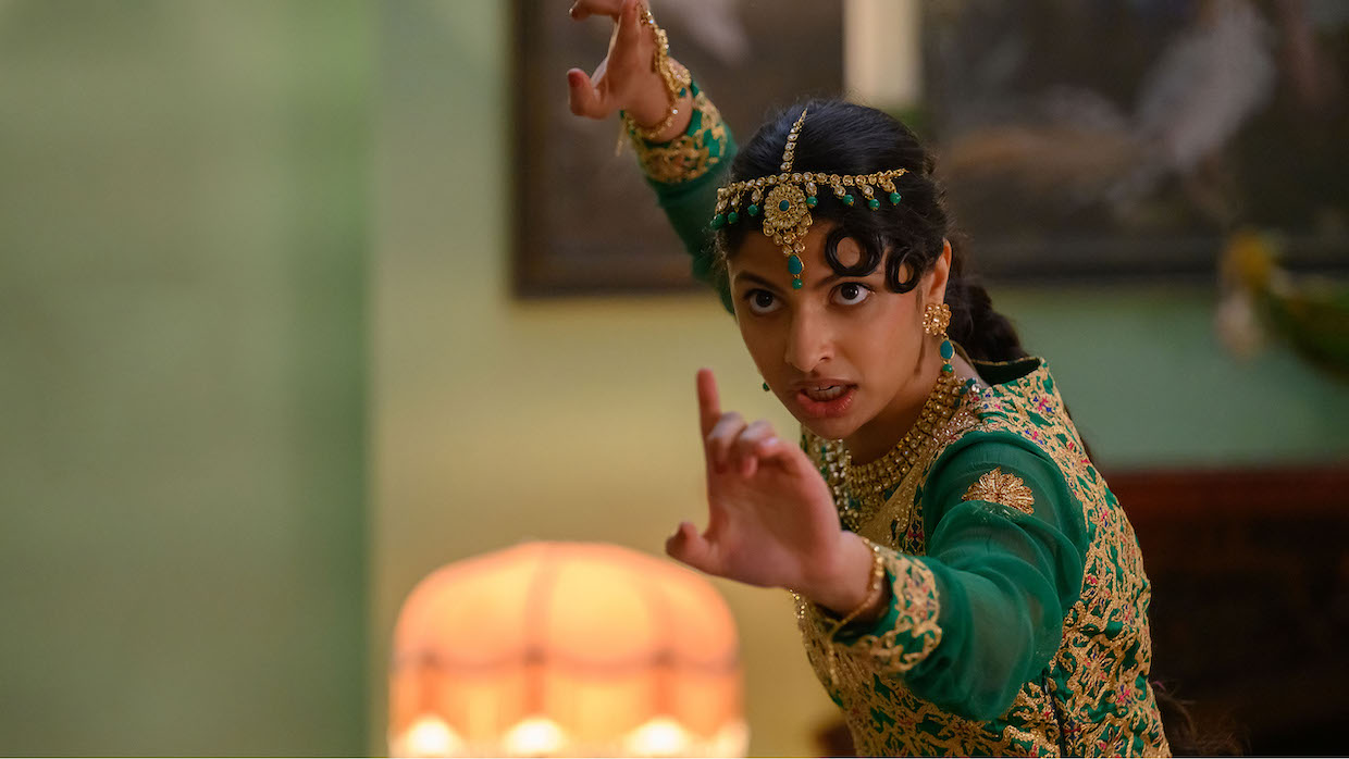 A young woman wears a green gown embroildered with gold details. She wears an ornate gold headpiece and holds her hands up in combat.