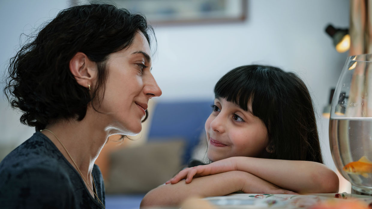 A woman with cropped, curly brown hair sits next to a young girl with straight black hair and bangs. They gaze at each other.