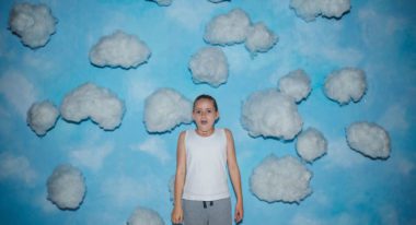 A young girl with blond hair wears a white tank top and gray sweatpants and stands in front of a wall with a blue sky and clouds painted on it.