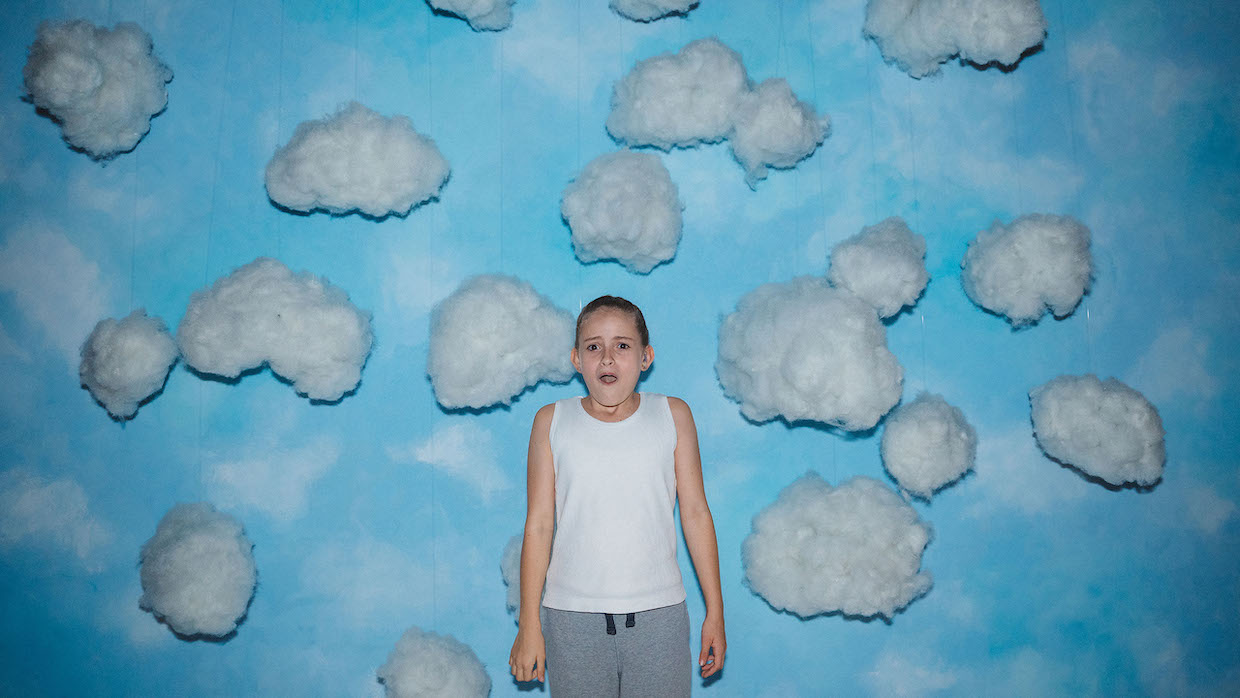 A young girl with blond hair wears a white tank top and gray sweatpants and stands in front of a wall with a blue sky and clouds painted on it.