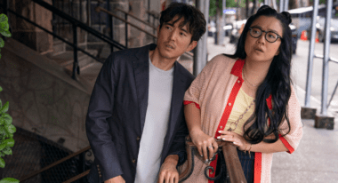 An Asian man and woman stand on the street, leaning slightly to their left while staring at something in the distance.