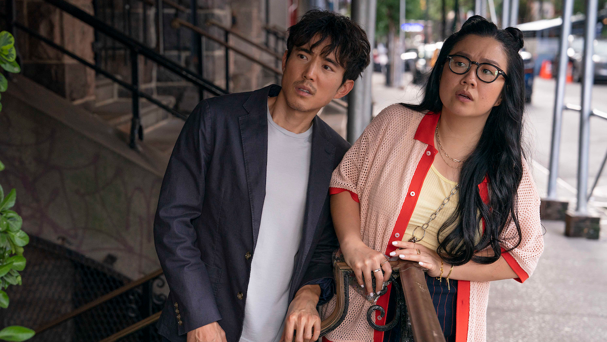 An Asian man and woman stand on the street, leaning slightly to their left while staring at something in the distance.