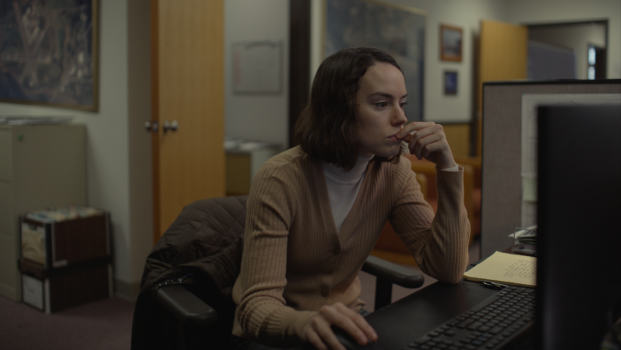 A woman with a shoulder-length bob haircut wears a camel-colored cardigan and sits in front of a computer at a desk in an office. She touches her fingers to her lips absentmindedly.