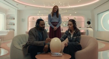 A young couple sits in a retro-futuristic room with pink-hued underlights. An egg-shaped object, perhaps really an egg, sits on a small wooden table in front of them. Behind them, a woman with shoulder-length blond hair looks over their shoulders.