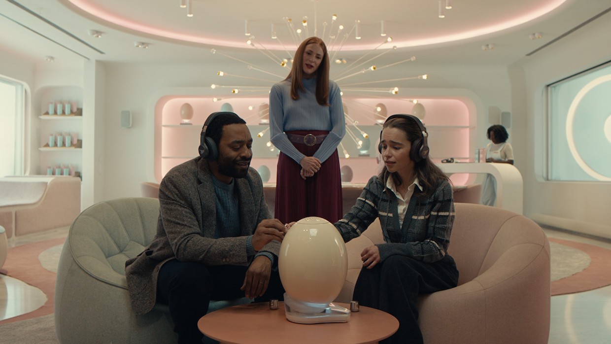 A young couple sits in a retro-futuristic room with pink-hued underlights. An egg-shaped object, perhaps really an egg, sits on a small wooden table in front of them. Behind them, a woman with shoulder-length blond hair looks over their shoulders.