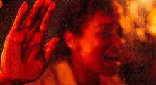 A woman screams with her hand pressed against a window. She is bathed in deep red light.