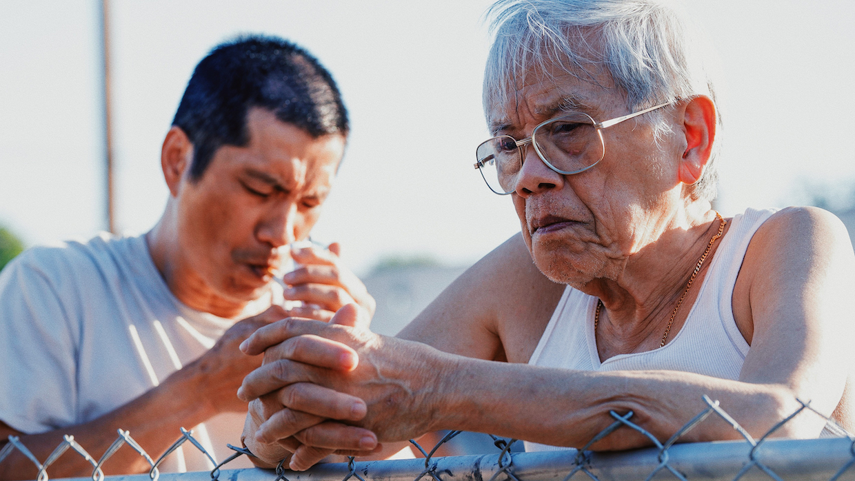 A young man wearing a gray t-shirt smokes a cigarette while an elderly man in a white tank top leans on a chainlink fence behind him.