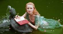 A woman with long, curly blond hair floats next to a metal turtle fountain in a body of water. She wears an ornate green dress.