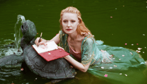 A woman with long, curly blond hair floats next to a metal turtle fountain in a body of water. She wears an ornate green dress.