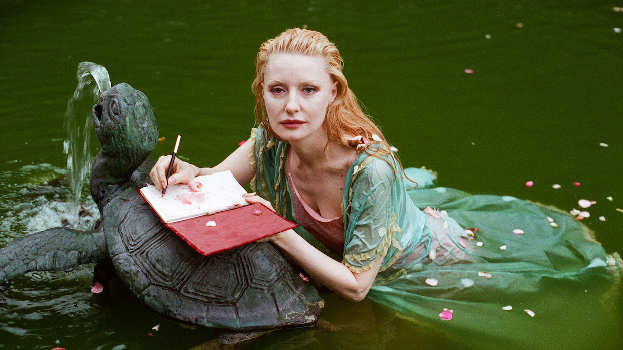 A woman with long, curly blond hair floats next to a metal turtle fountain in a body of water. She wears an ornate green dress.