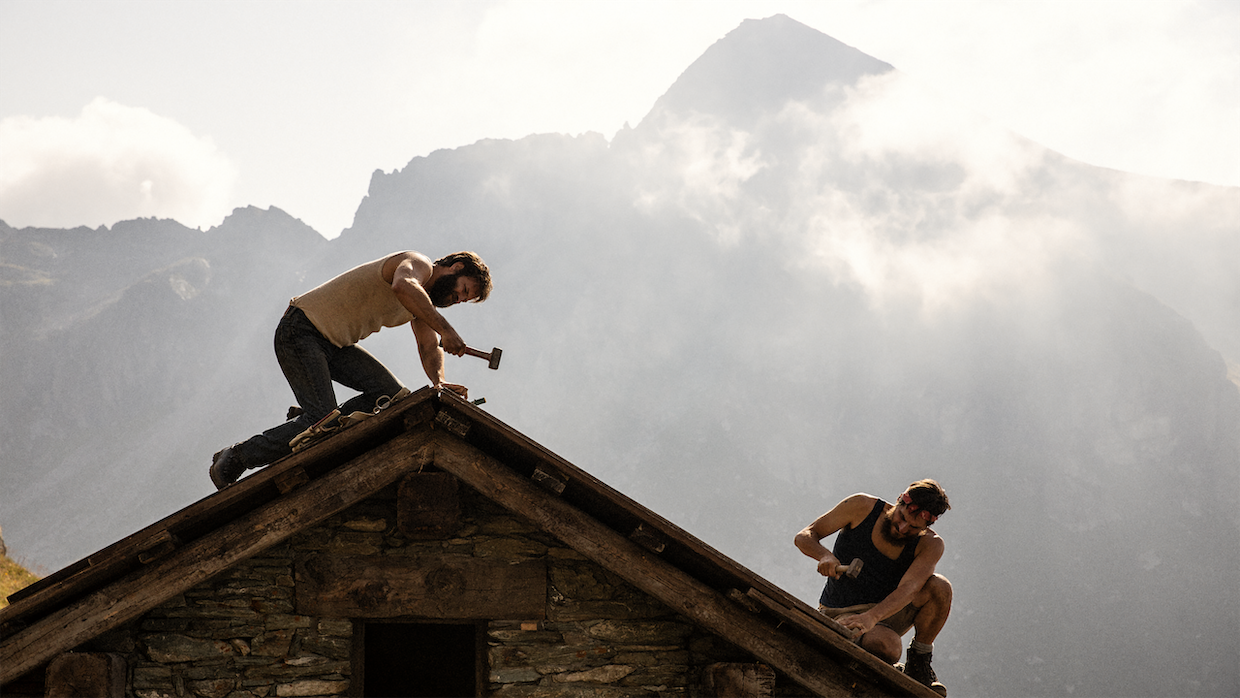 Two men work on a house's roof with hammers with the mountains of the Swiss Alps in the background.