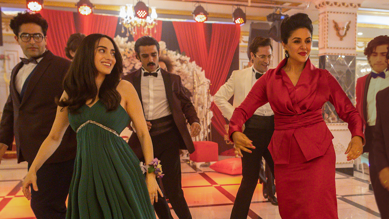 An Iranian-American family lines up on a dance floor. A mother and daughter stand in front of male relatives wearing tuxedos. The younger woman wears a green gown while the mother wears a red suit skirt ensemble.