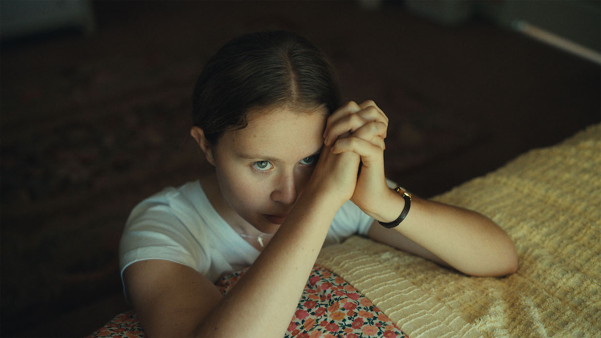 A young girl with brown hair tied back in a low ponytail kneels beside her bed and folds her hands in prayer. Her eyes are open and she looks up.