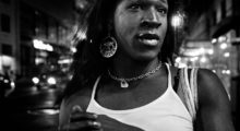 A Black trans woman wears a white tank top, a chain necklace and large hoop earrings that say "Taurus" with a picture of a bull. She walks in NYC's meatpacking district behind her.